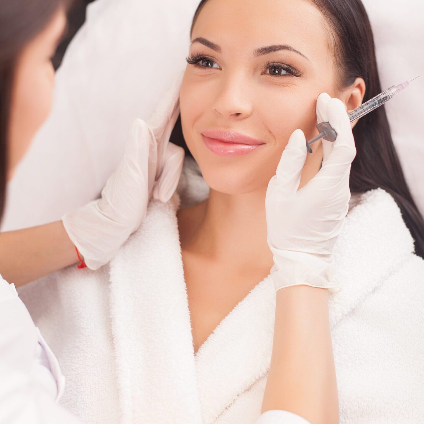 Beautiful young woman is getting botox injection in her face. She is sitting and smiling. The female doctor is checking her skin with concentration and holding syringe
